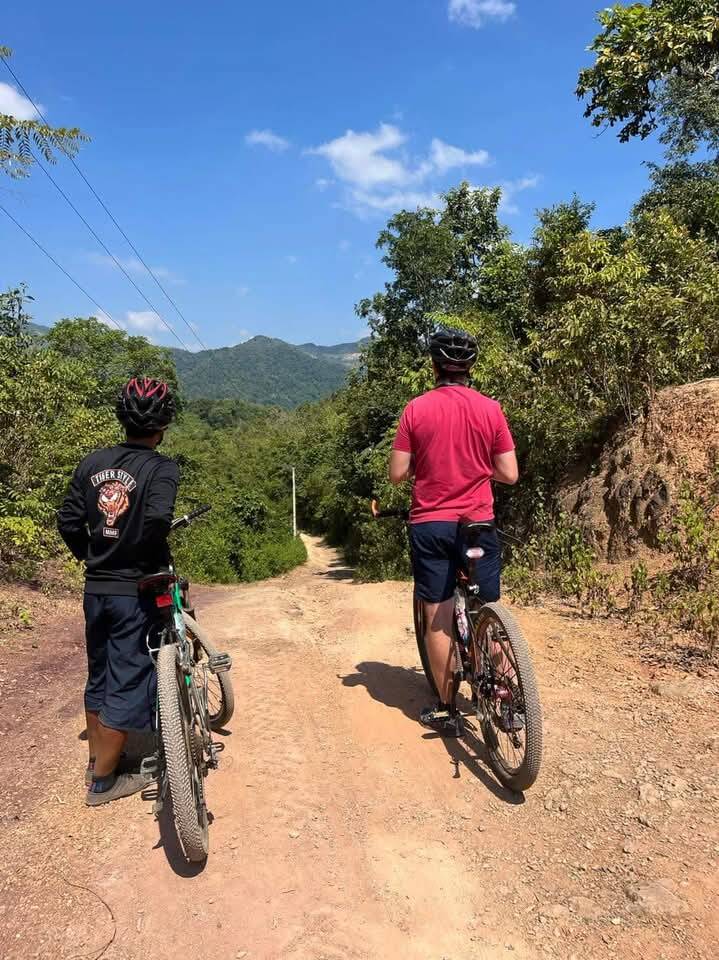 Two people with bikes on a dirt road, mountains ahead
